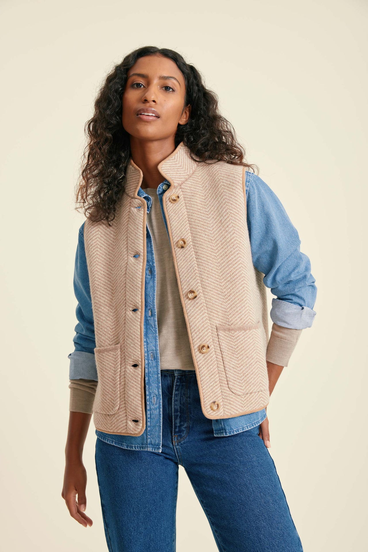 Woman wearing a beige vest over a blue shirt and jeans on a beige background