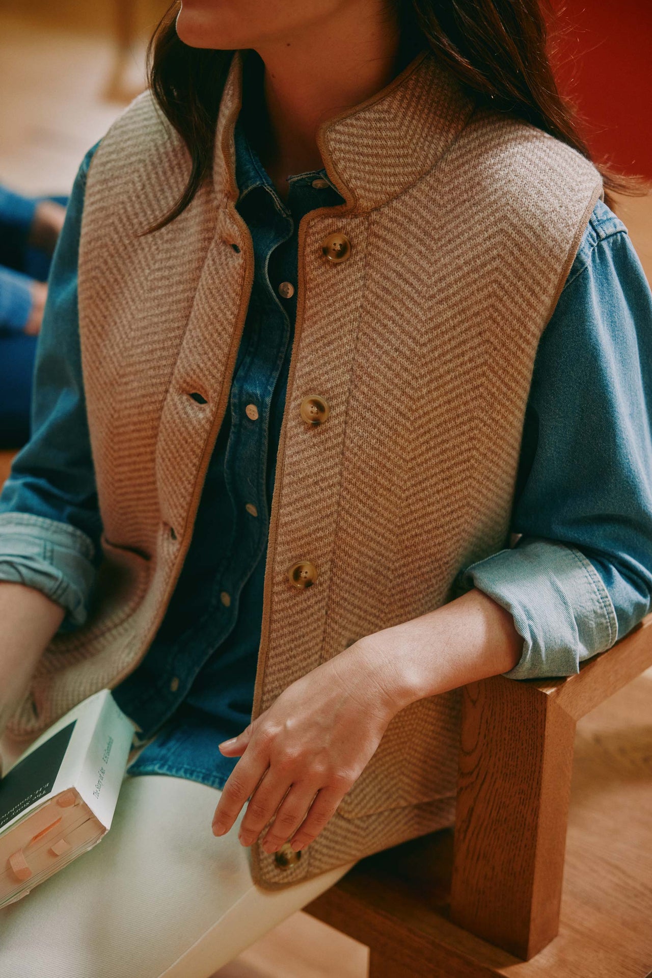 Person wearing a brown herringbone vest over a blue shirt, sitting on a wooden stool.