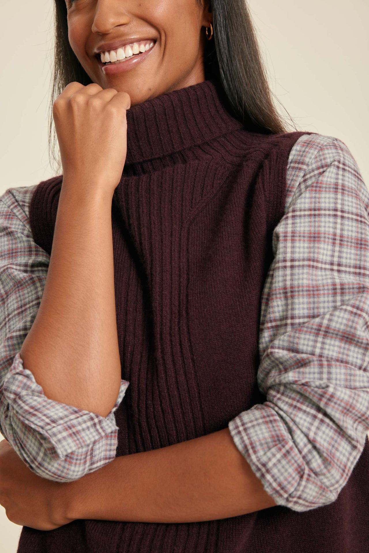 Woman wearing a dark brown ribbed vest over a plaid shirt against a beige background