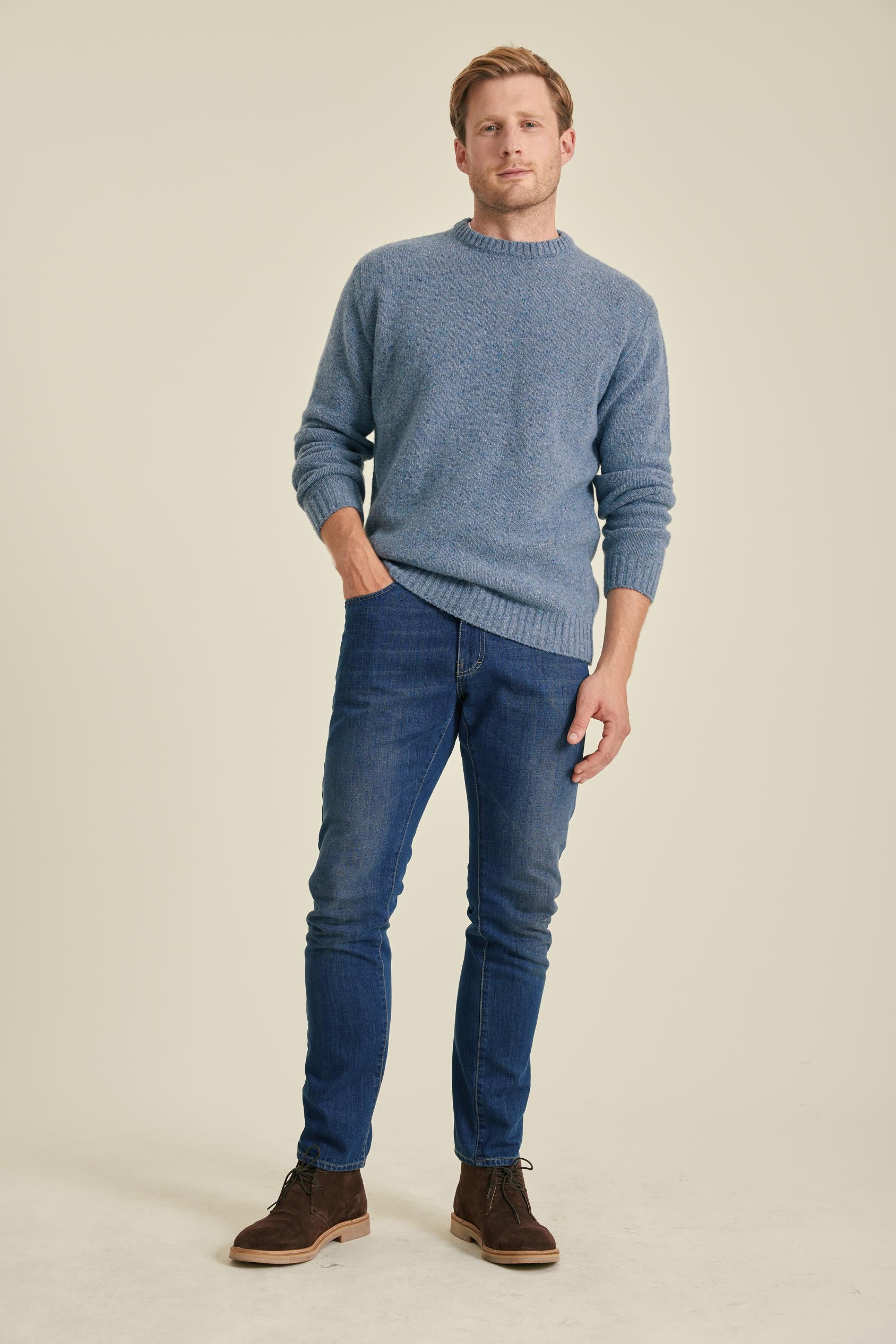 Man wearing a blue sweater and jeans standing against a beige background
