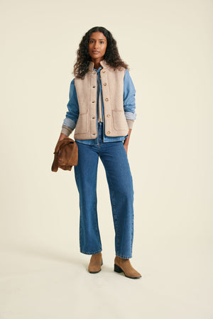 Woman wearing a beige vest over a blue shirt and jeans, holding a brown leather bag on a beige background