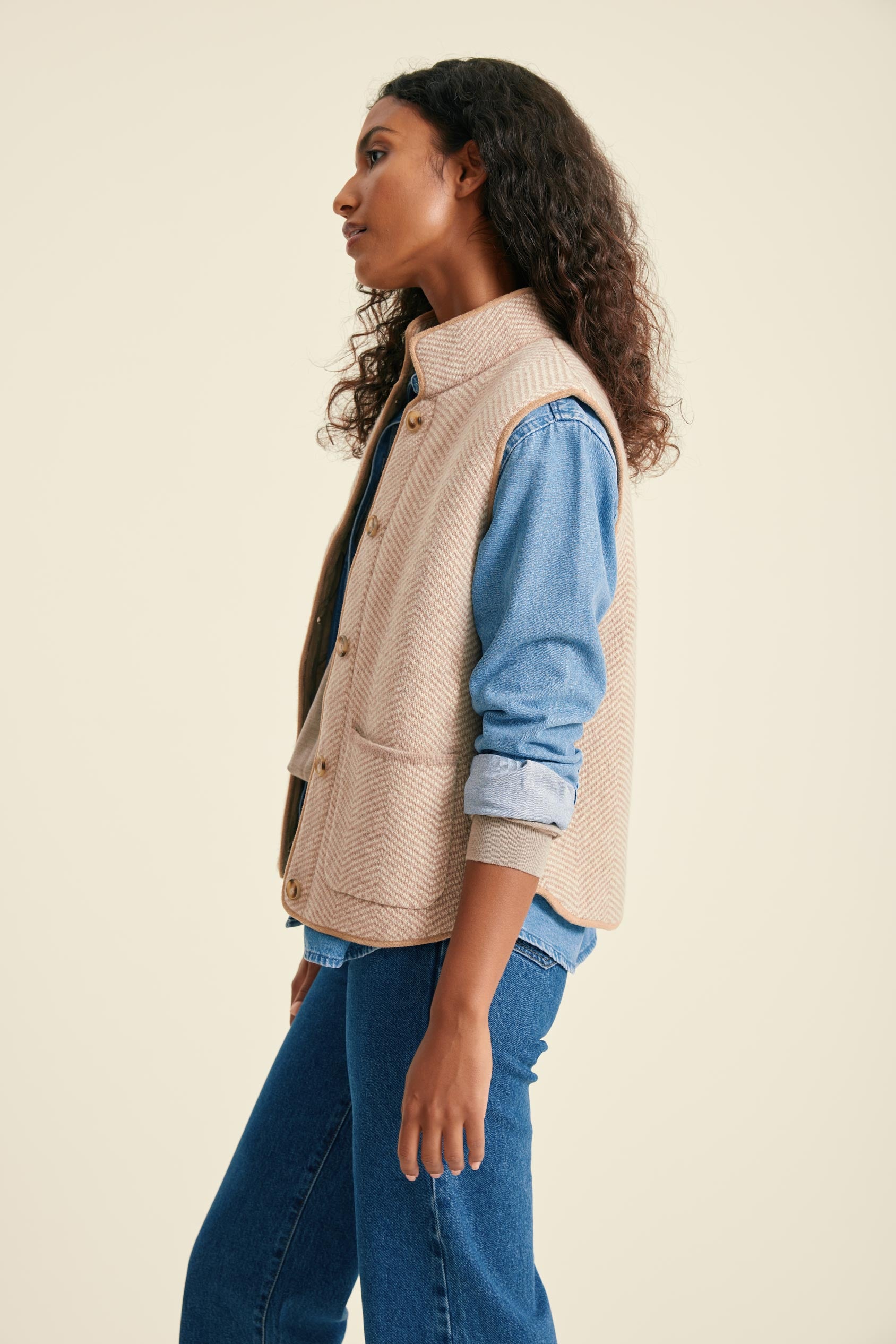 Woman wearing a beige vest over a blue shirt and jeans on a beige background