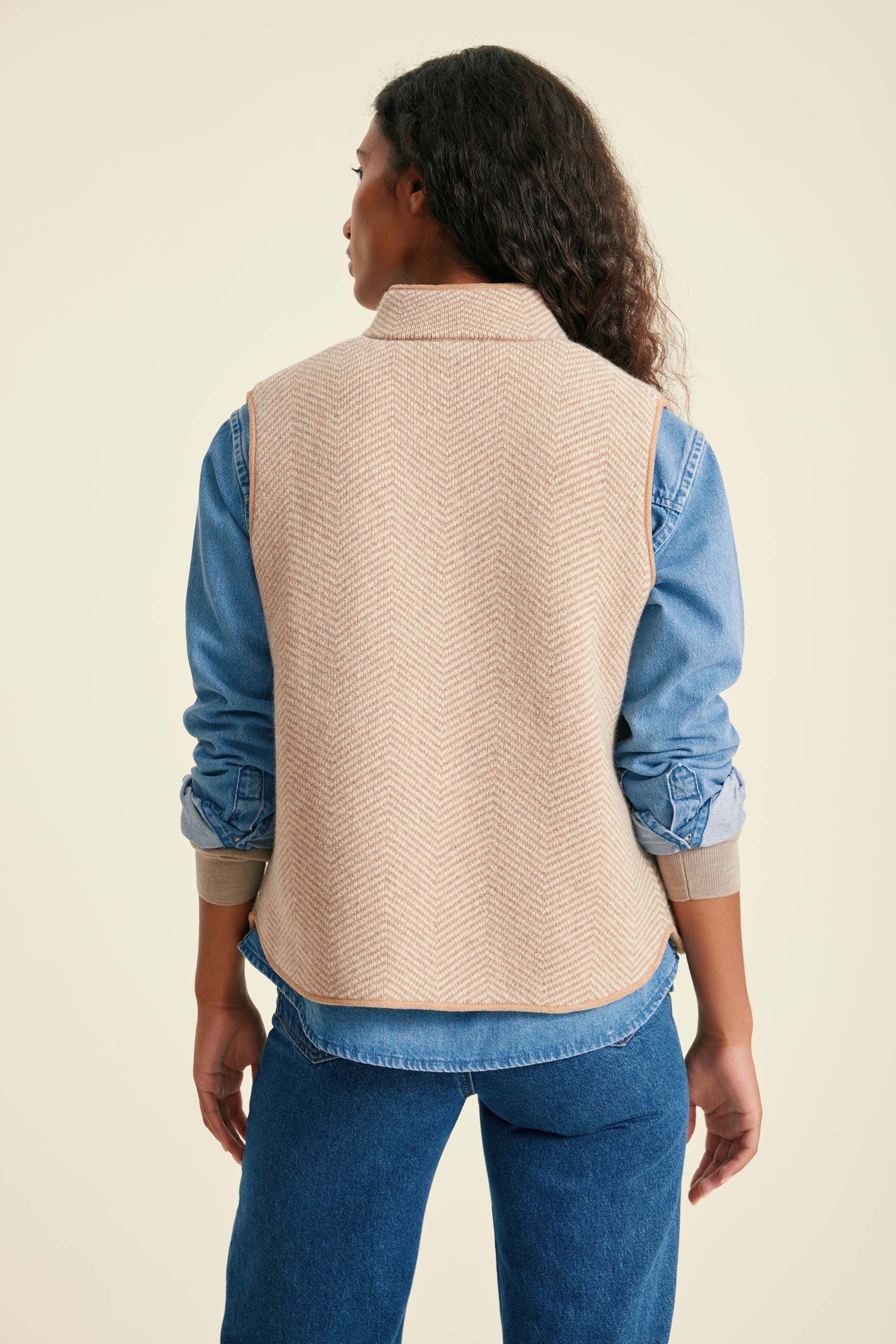 Person wearing a beige vest over a blue shirt and jeans on a beige background
