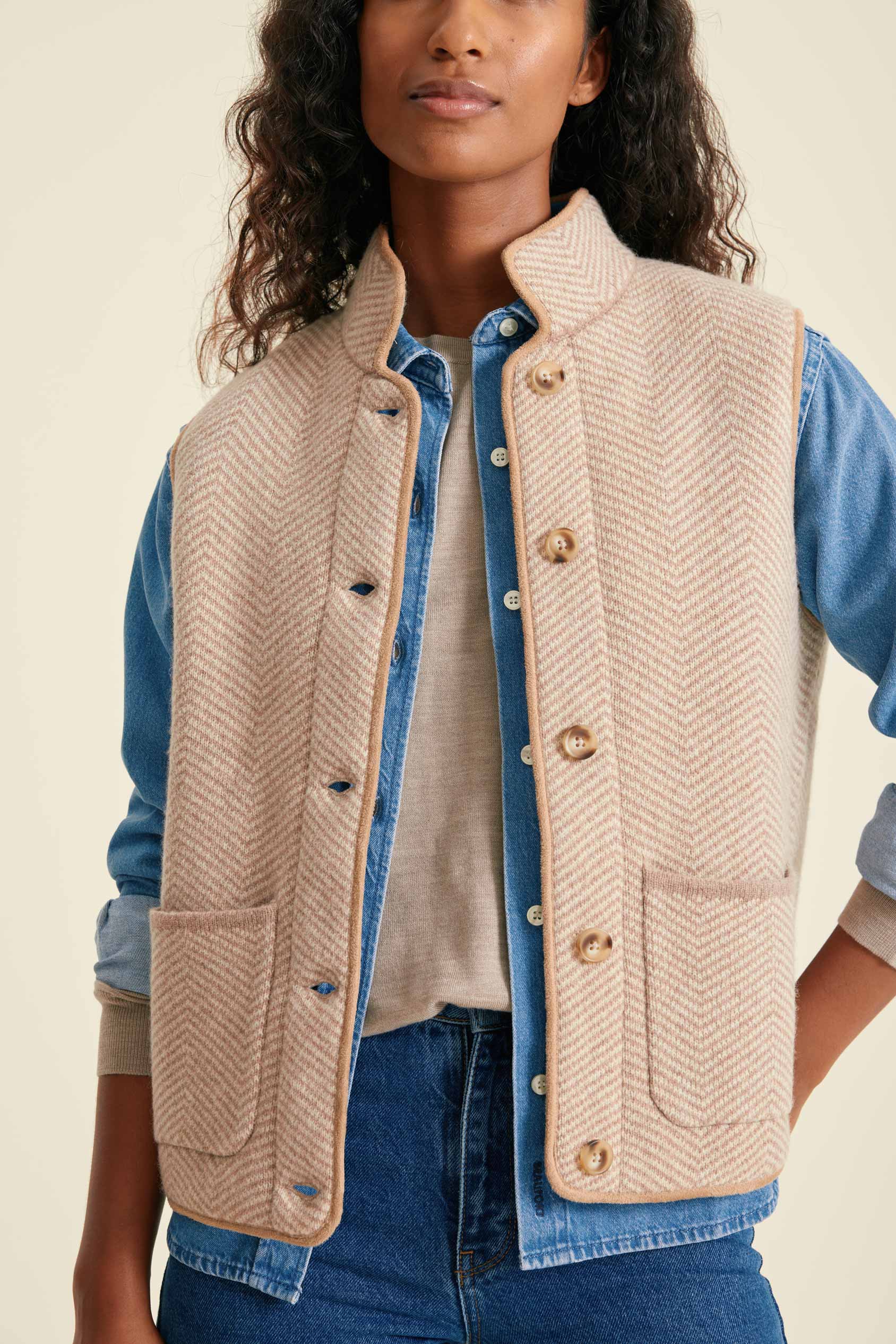 Woman wearing a beige herringbone vest over a blue shirt and jeans on a beige background