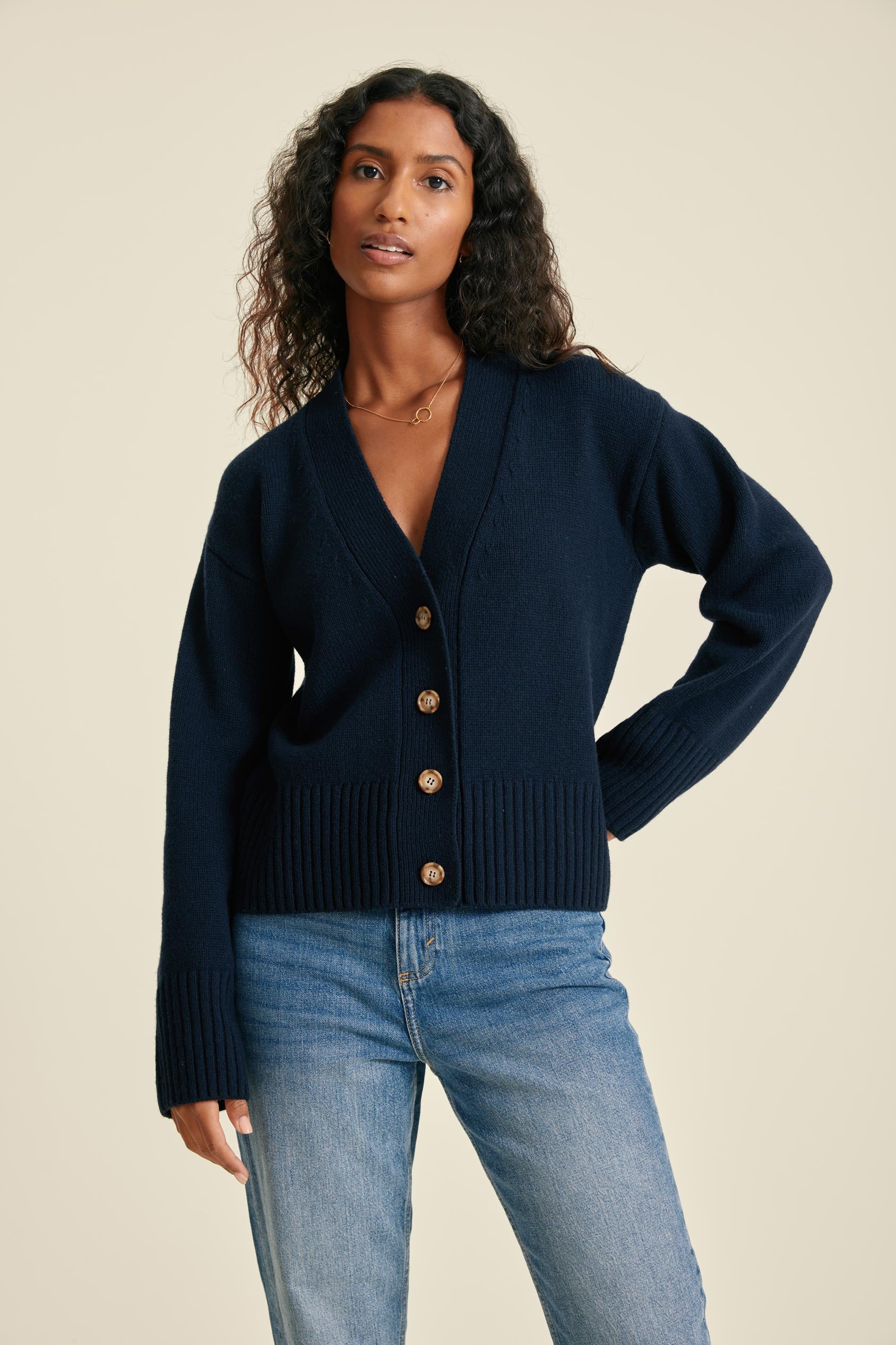 Woman wearing a navy cardigan with gold buttons and blue jeans on a beige background