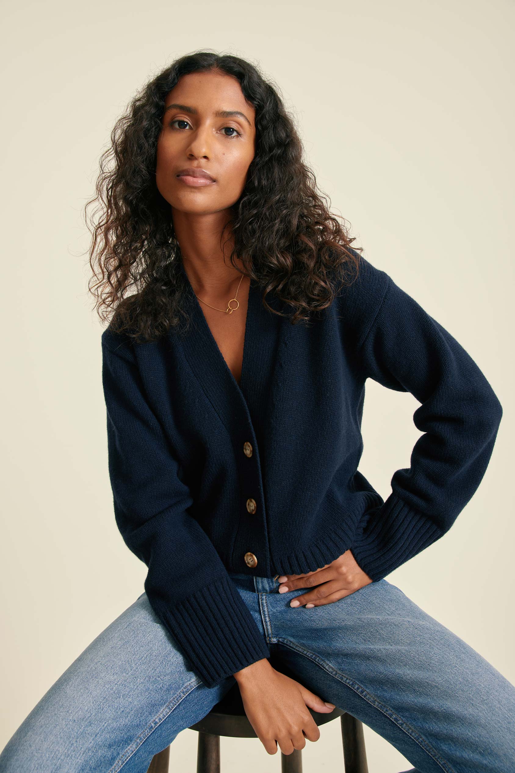 Woman wearing a navy cardigan and blue jeans sitting on a stool against a beige background