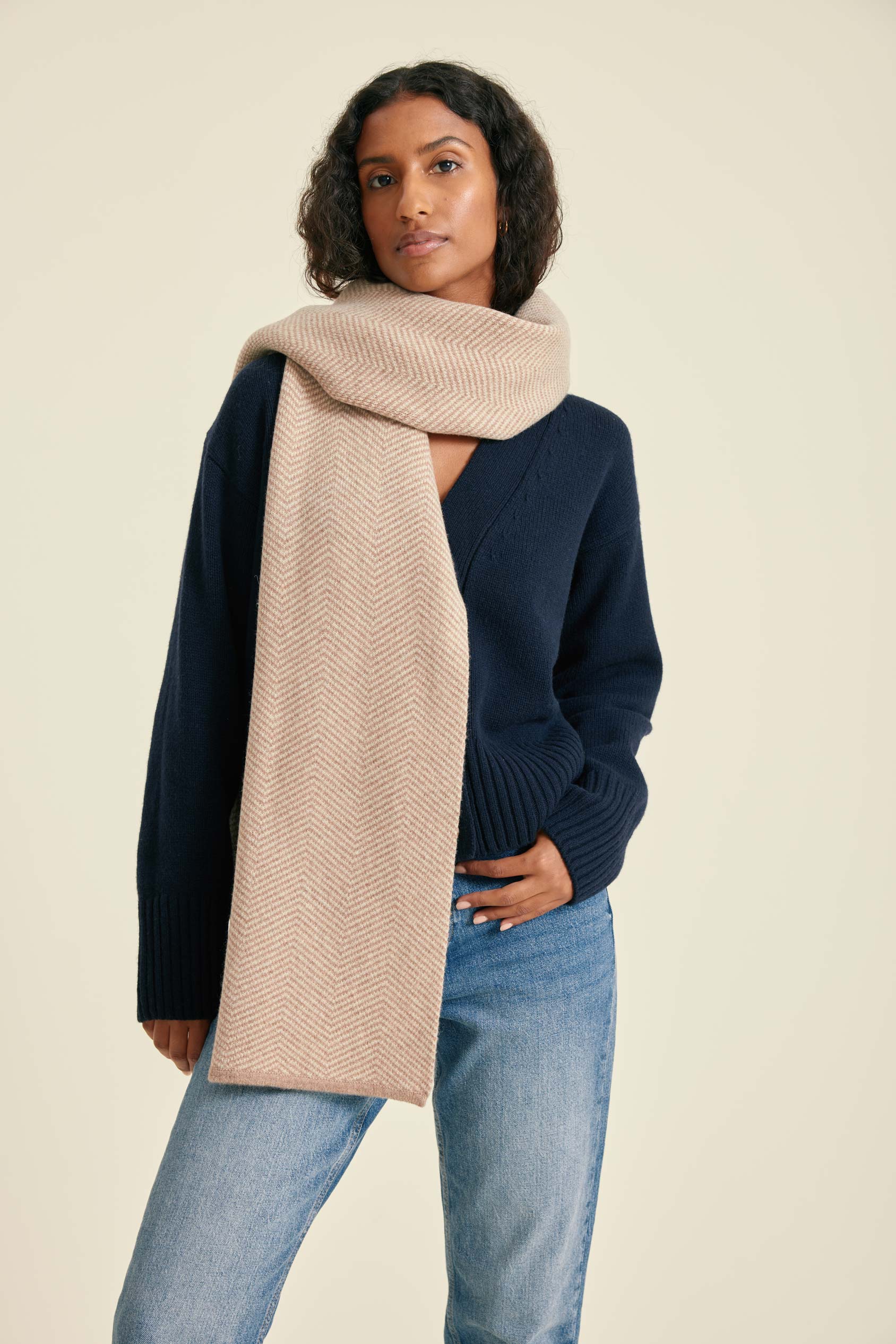 Woman wearing a beige scarf, navy sweater, and blue jeans on a beige background