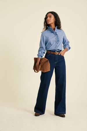 Woman wearing a blue shirt and dark blue jeans holding a brown bag on a beige background