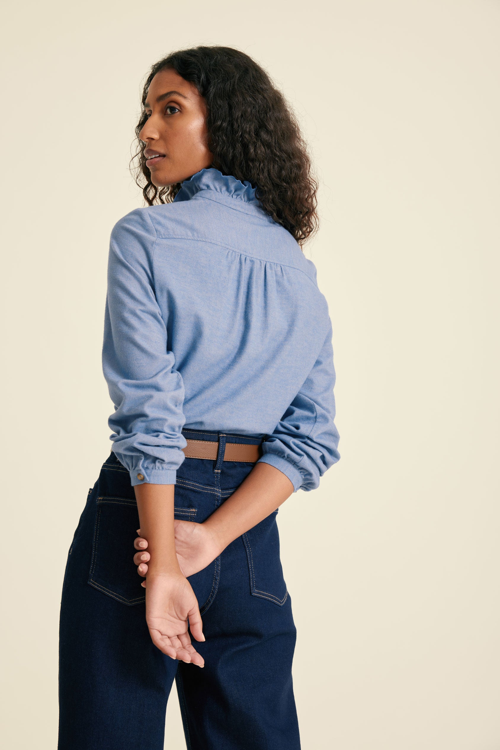 Woman wearing a blue denim shirt and dark jeans against a beige background