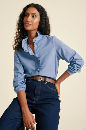 Woman wearing a blue shirt with ruffled collar and dark jeans on a beige background