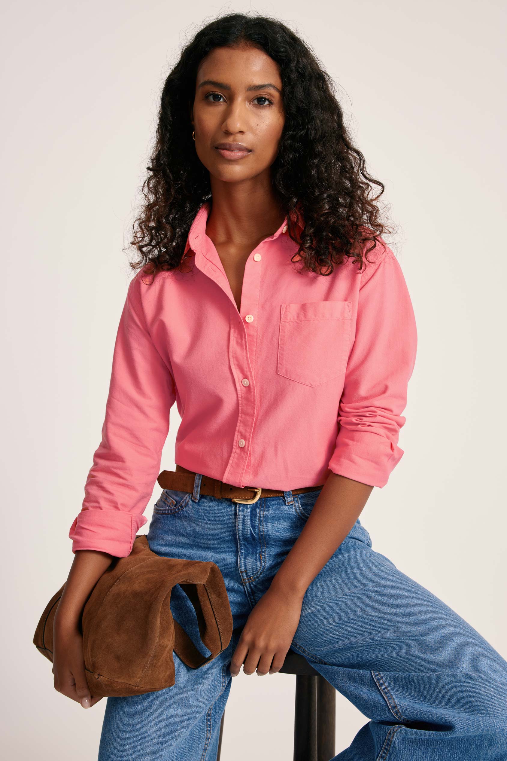 Woman wearing a pink shirt and blue jeans, holding a brown bag against a white background