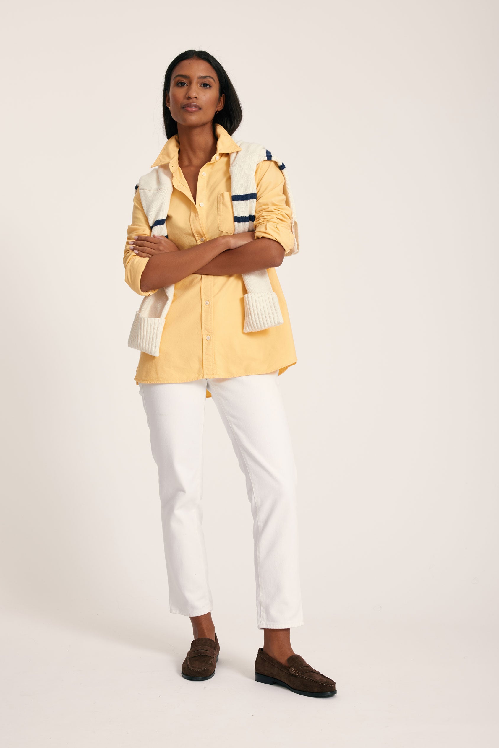 Woman wearing a yellow jacket with a white and blue patterned design, white pants, and brown shoes on a light background.