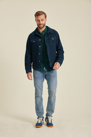 Man wearing a denim jacket, green shirt, and blue jeans on a beige background