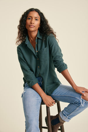 Woman wearing a green shirt and blue jeans sitting on a stool against a beige background