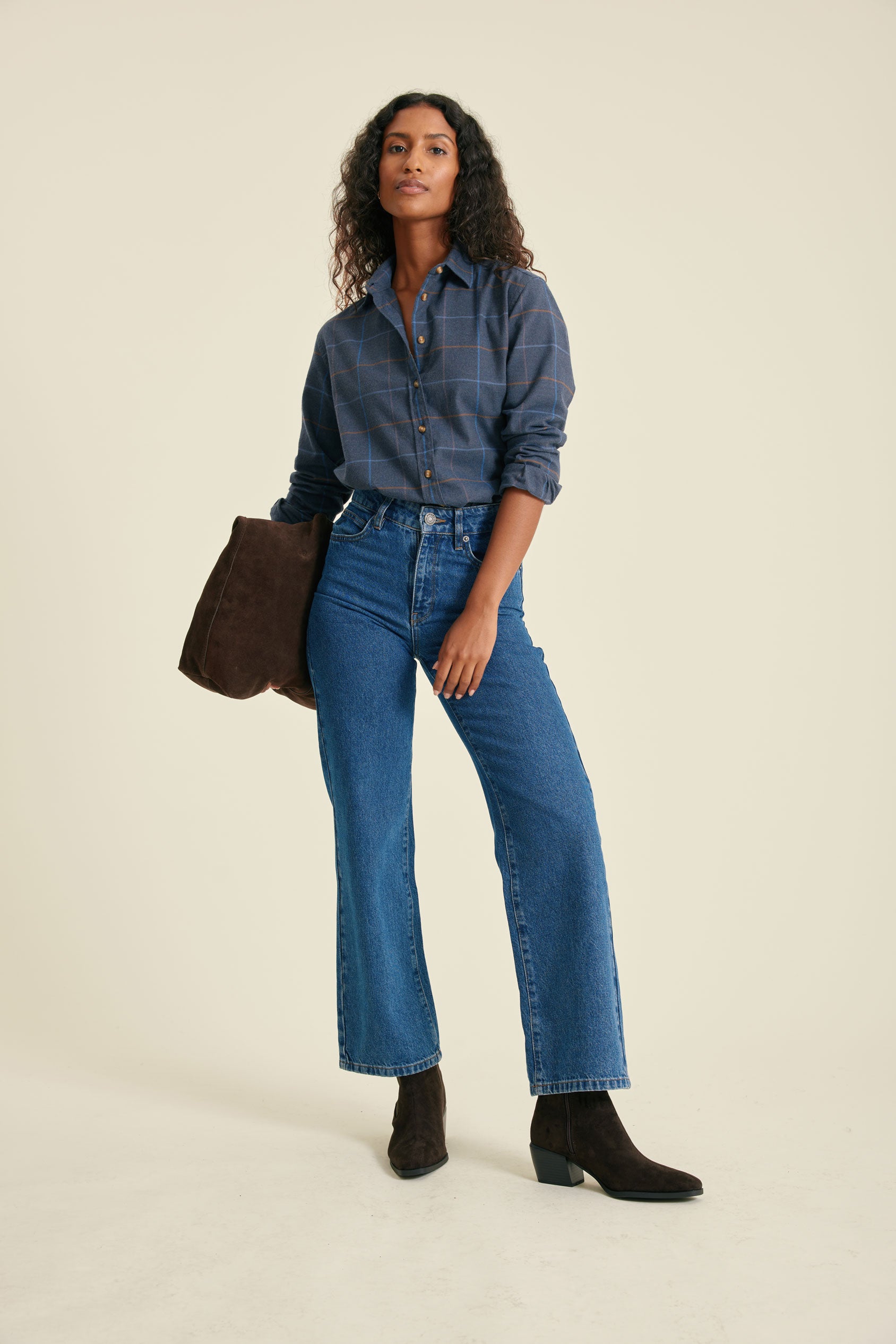Woman wearing a blue plaid shirt, blue jeans, and black boots, holding a brown leather bag on a beige background.