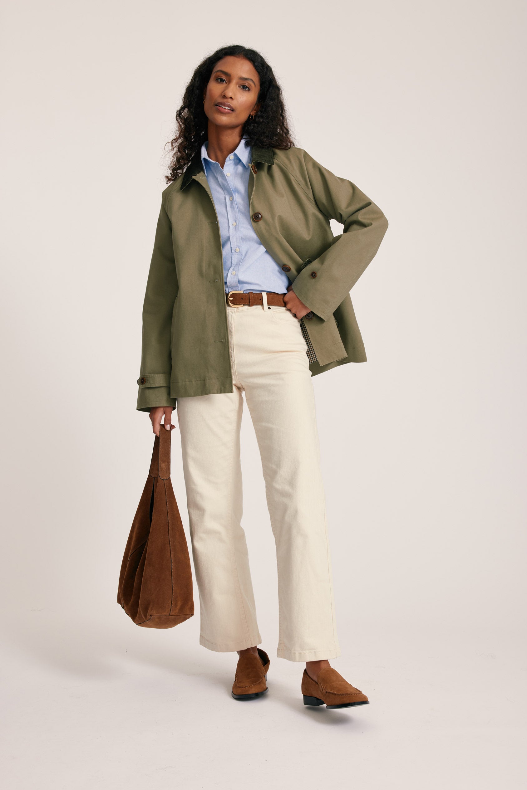 Woman wearing a green jacket, blue shirt, white pants, and brown shoes, holding a brown bag on a light background
