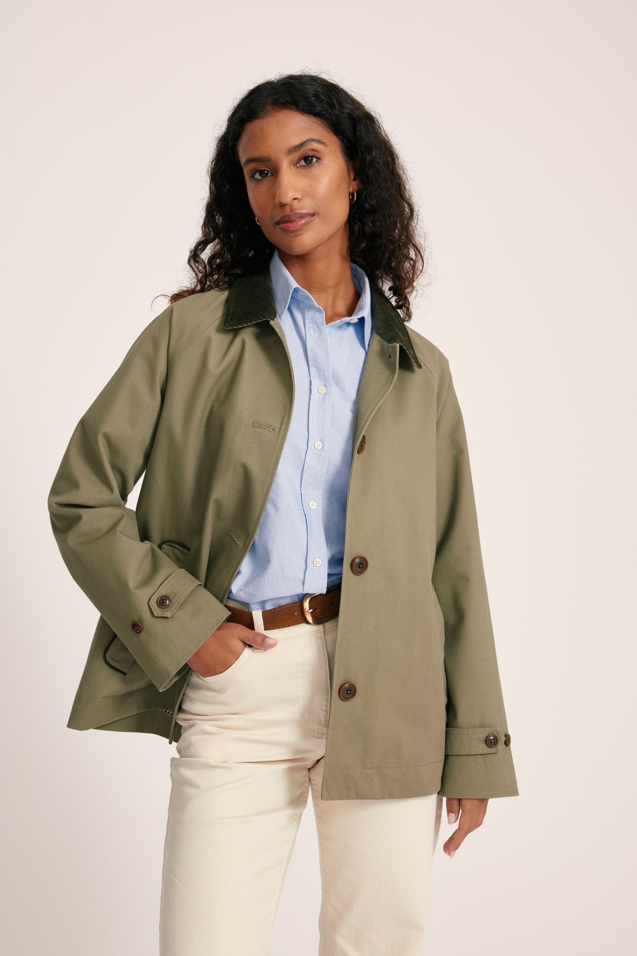 Woman wearing a green jacket over a blue shirt and white pants on a beige background