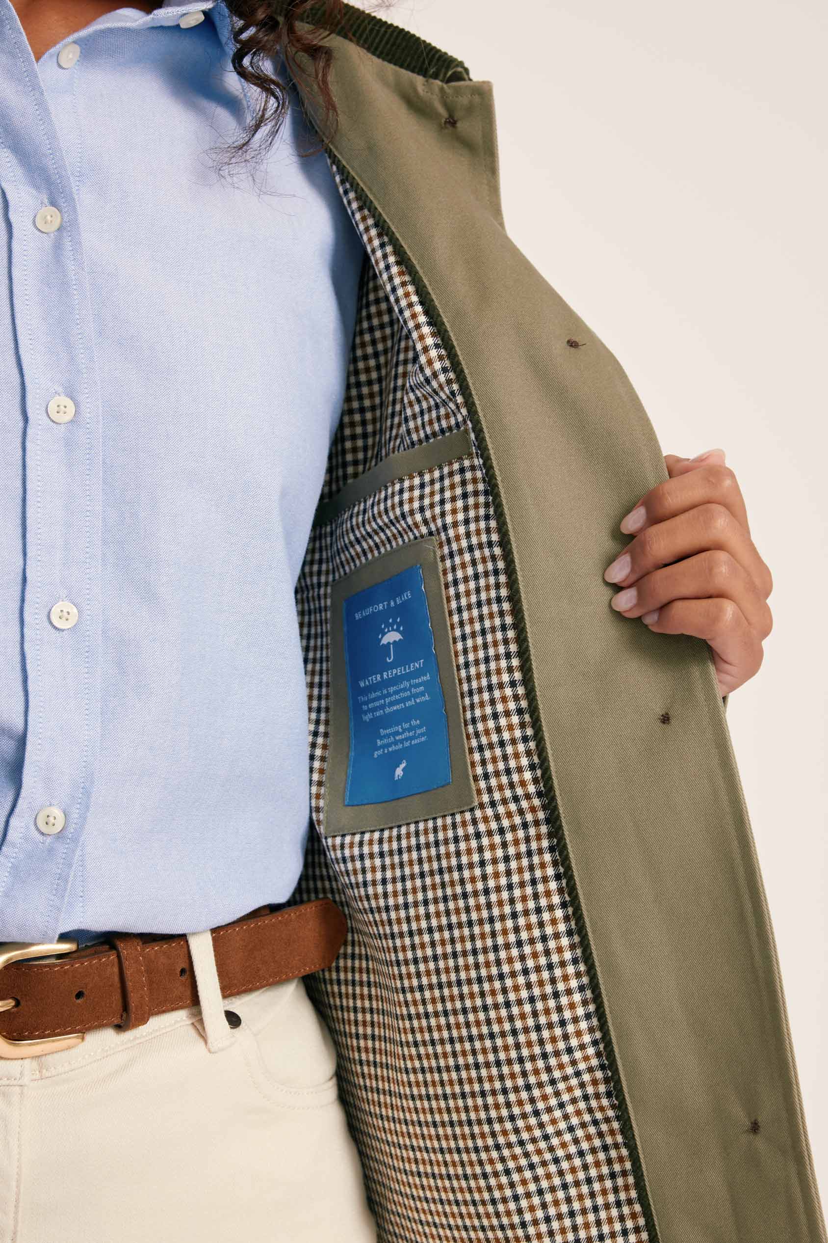 Person wearing a light blue shirt, beige pants, and a green coat with a checkered lining, holding the coat open to reveal the lining.