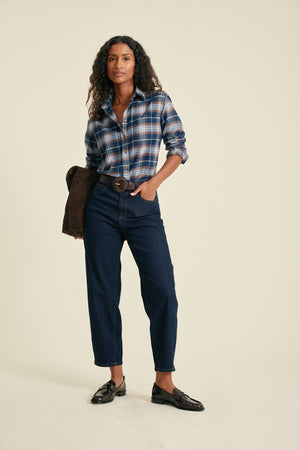 Woman wearing a plaid shirt, dark jeans, and black shoes on a beige background