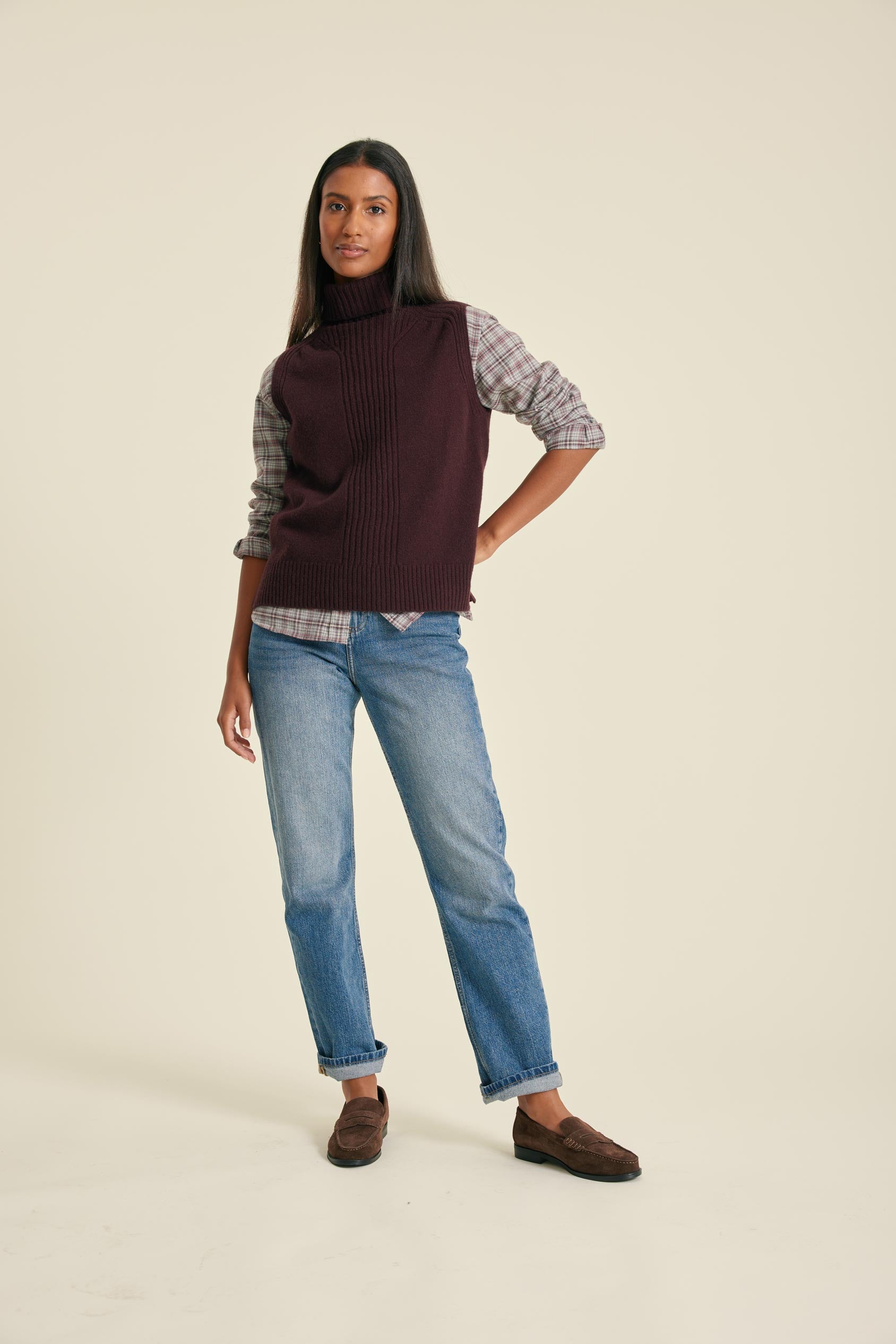Woman wearing a dark brown sweater over a plaid shirt with blue jeans on a beige background