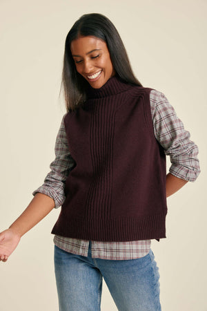 Woman wearing a dark brown knitted vest over a plaid shirt with a beige background