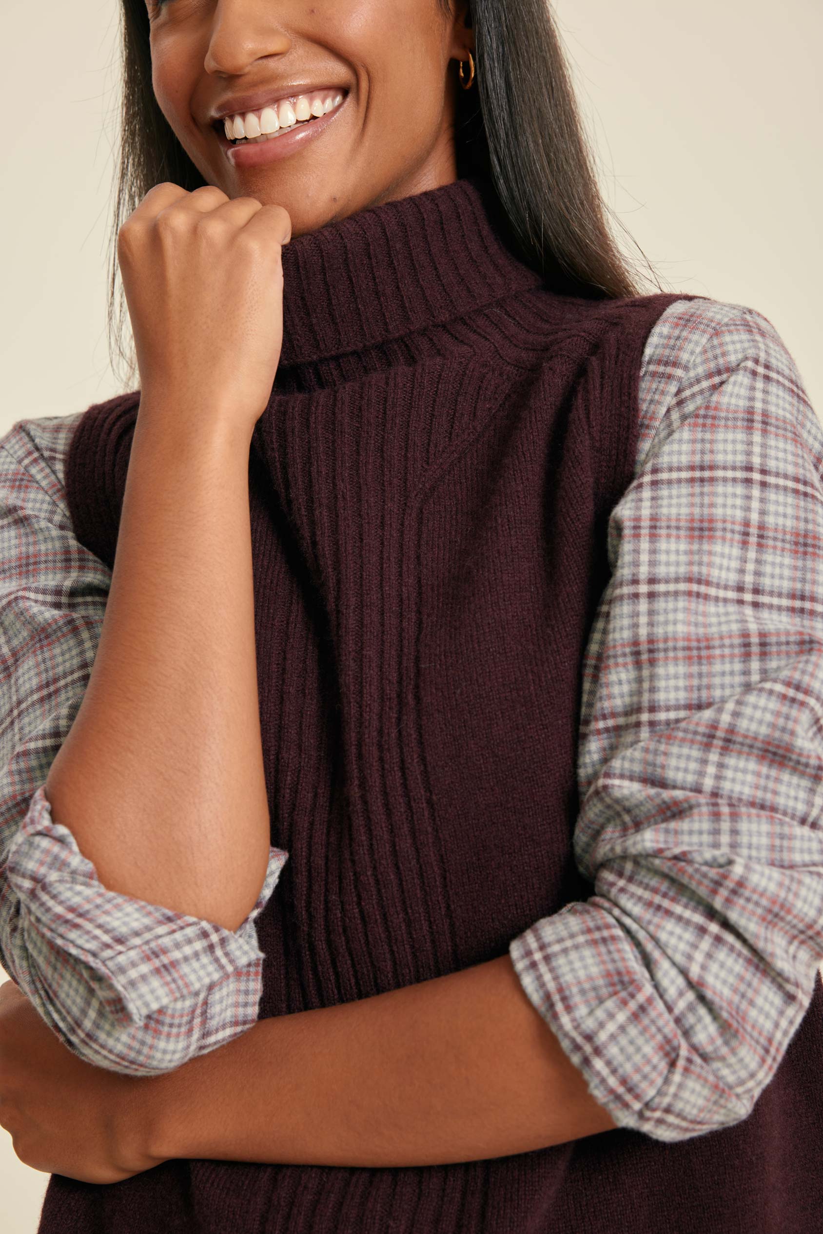 Woman wearing a dark brown ribbed vest over a plaid shirt against a beige background