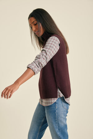 Woman wearing a maroon sweater vest over a plaid shirt with blue jeans on a beige background