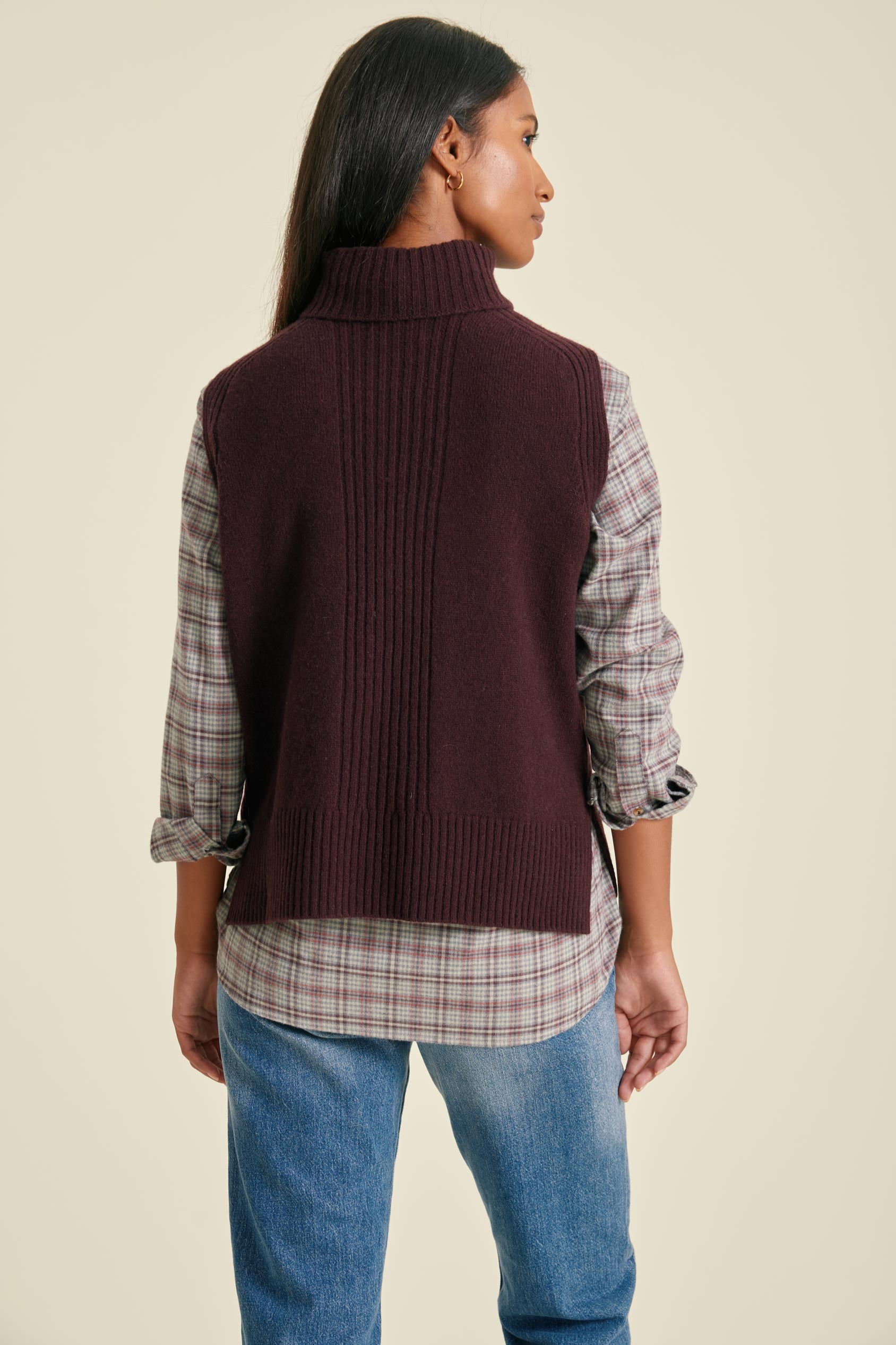Person wearing a brown vest over a plaid shirt with blue jeans on a beige background
