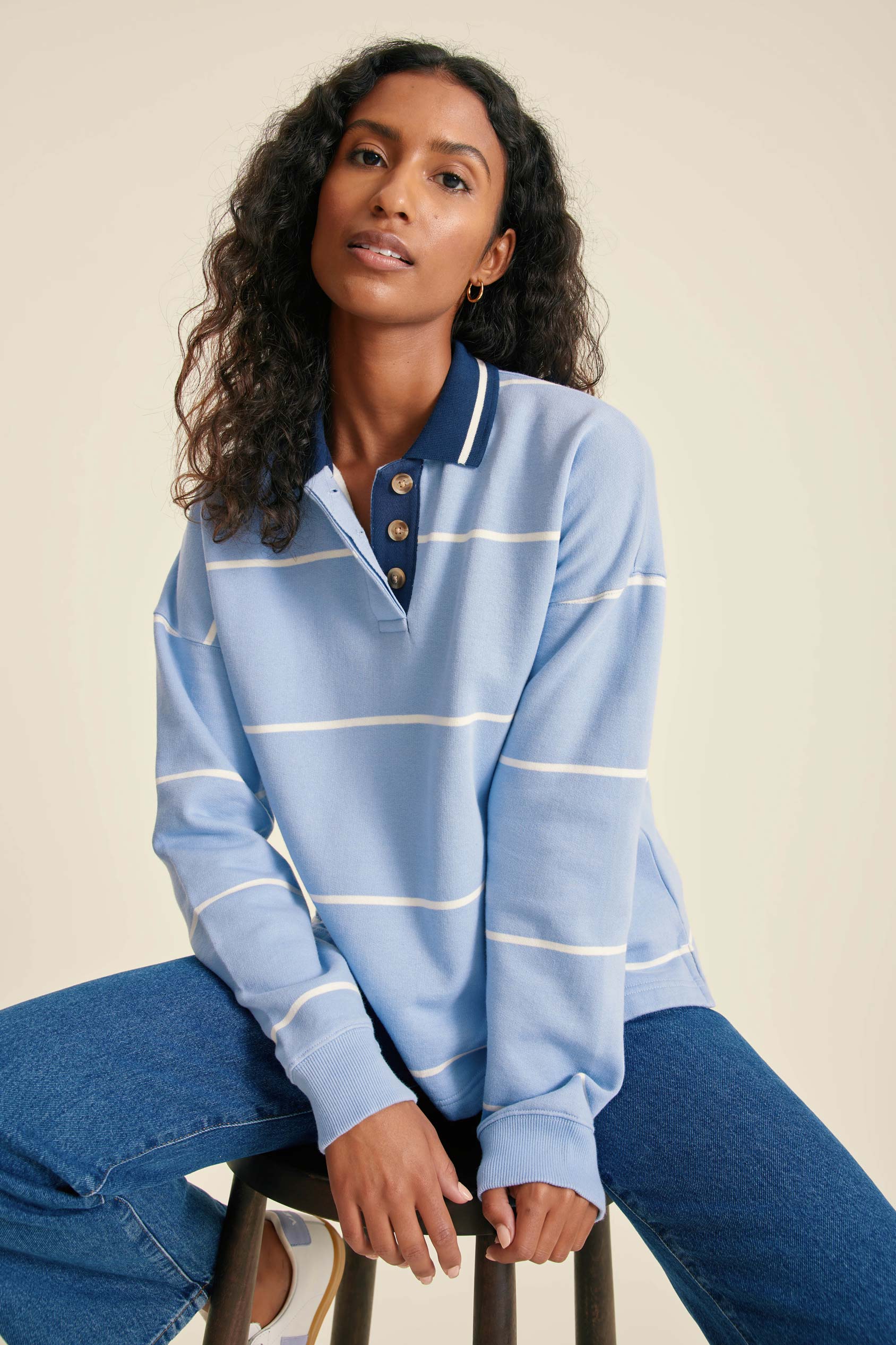 Woman wearing a blue striped sweater and jeans sitting on a stool against a beige background