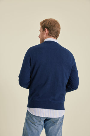 Man wearing a navy sweater over a white shirt with blue jeans on a beige background