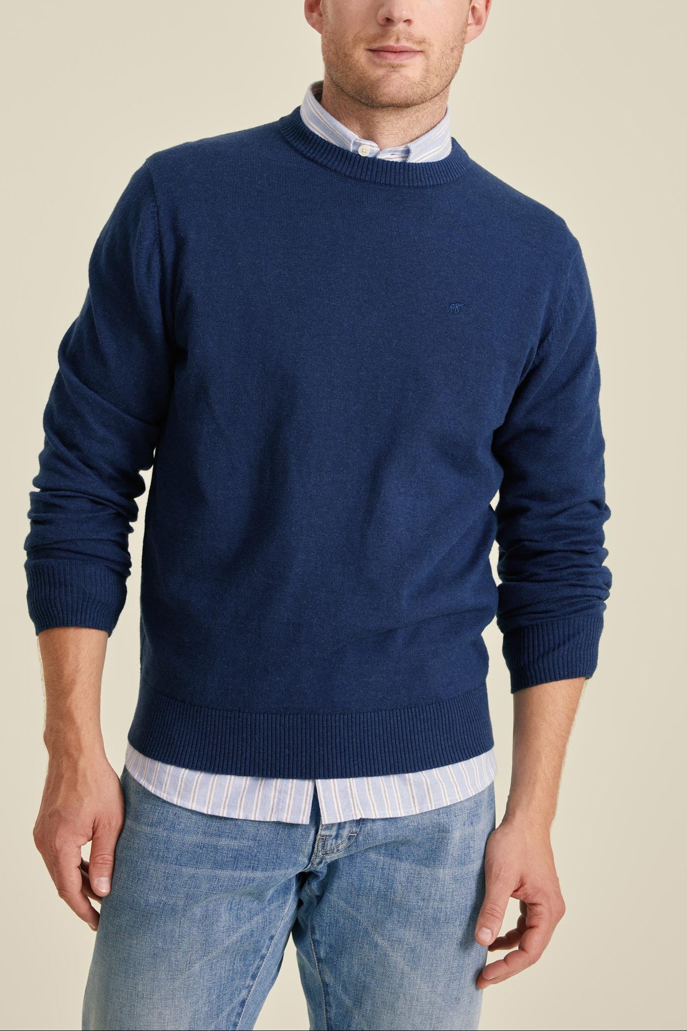 Man wearing a blue sweater over a collared shirt with light blue jeans on a beige background