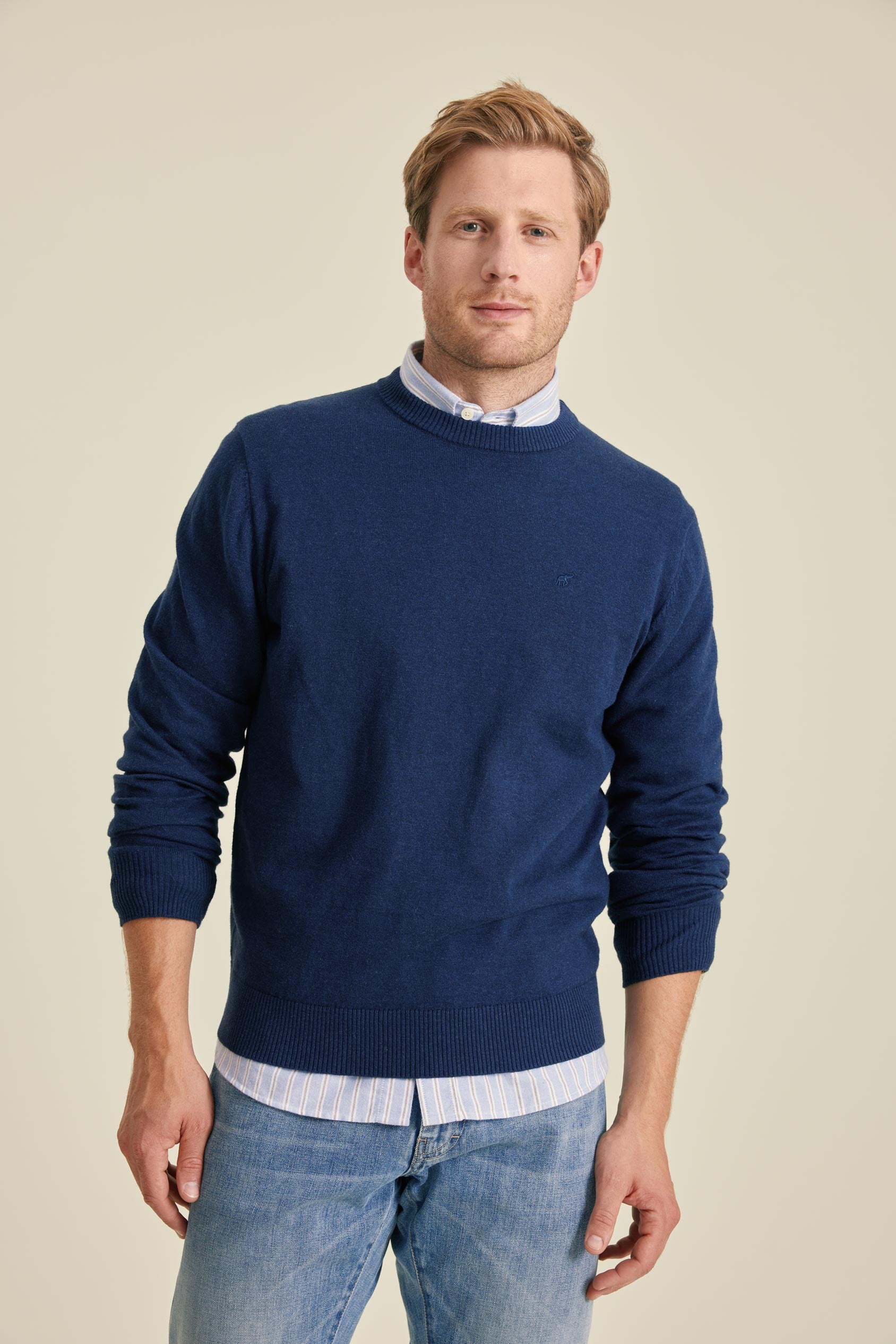 Man wearing a blue sweater over a collared shirt with light blue jeans on a beige background