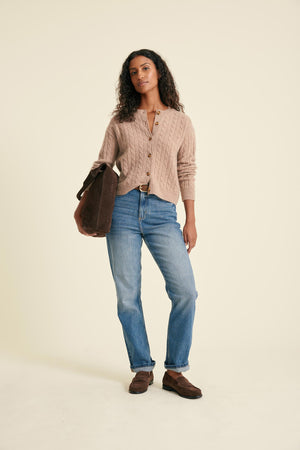 Woman wearing a beige cardigan, blue jeans, and brown shoes, holding a brown bag on a beige background