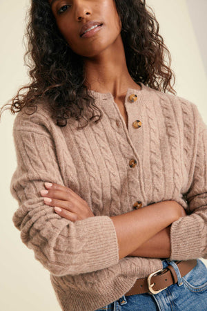 Woman wearing a beige cable knit cardigan with gold buttons on a plain background