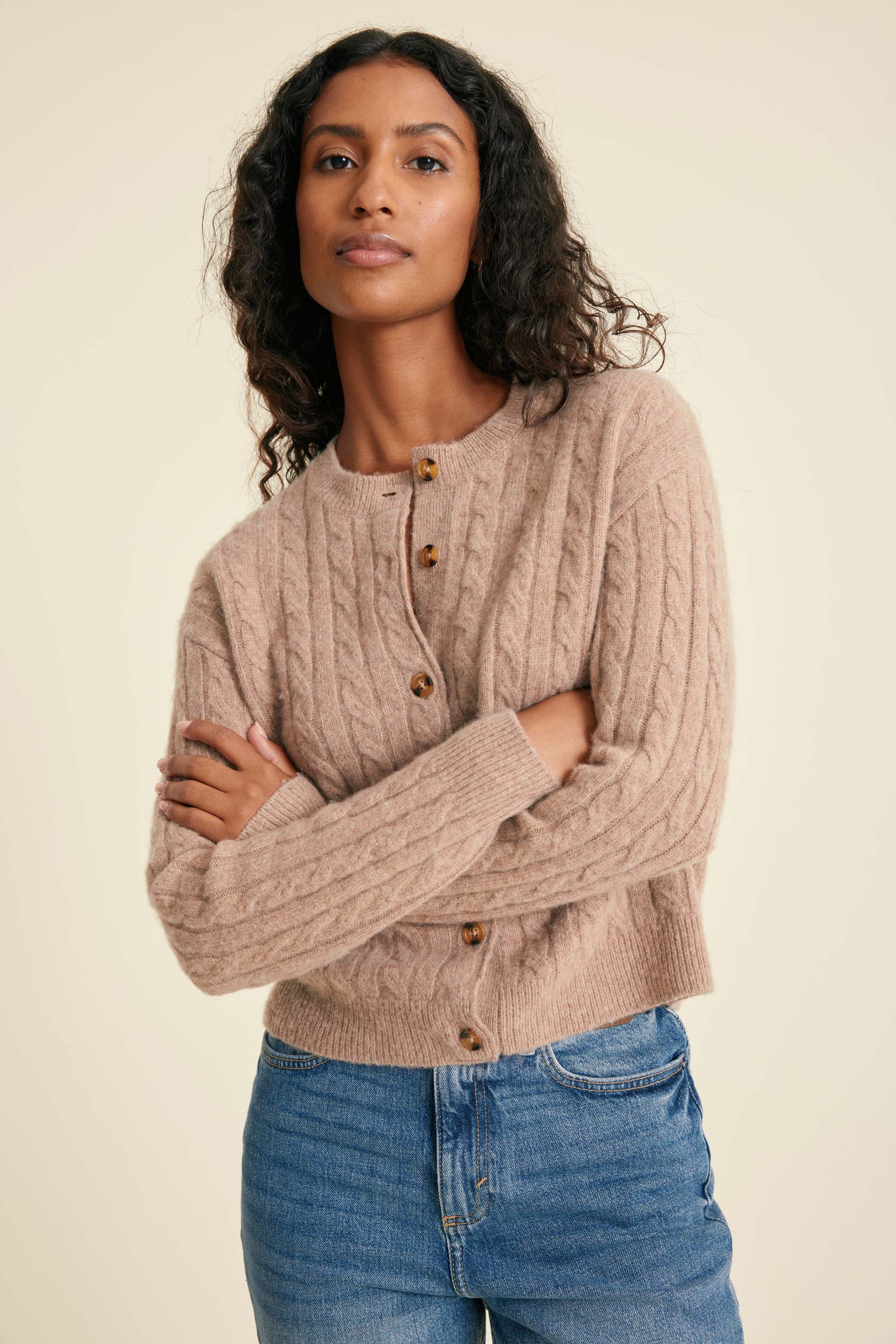 Woman wearing a beige cable knit cardigan and blue jeans against a beige background