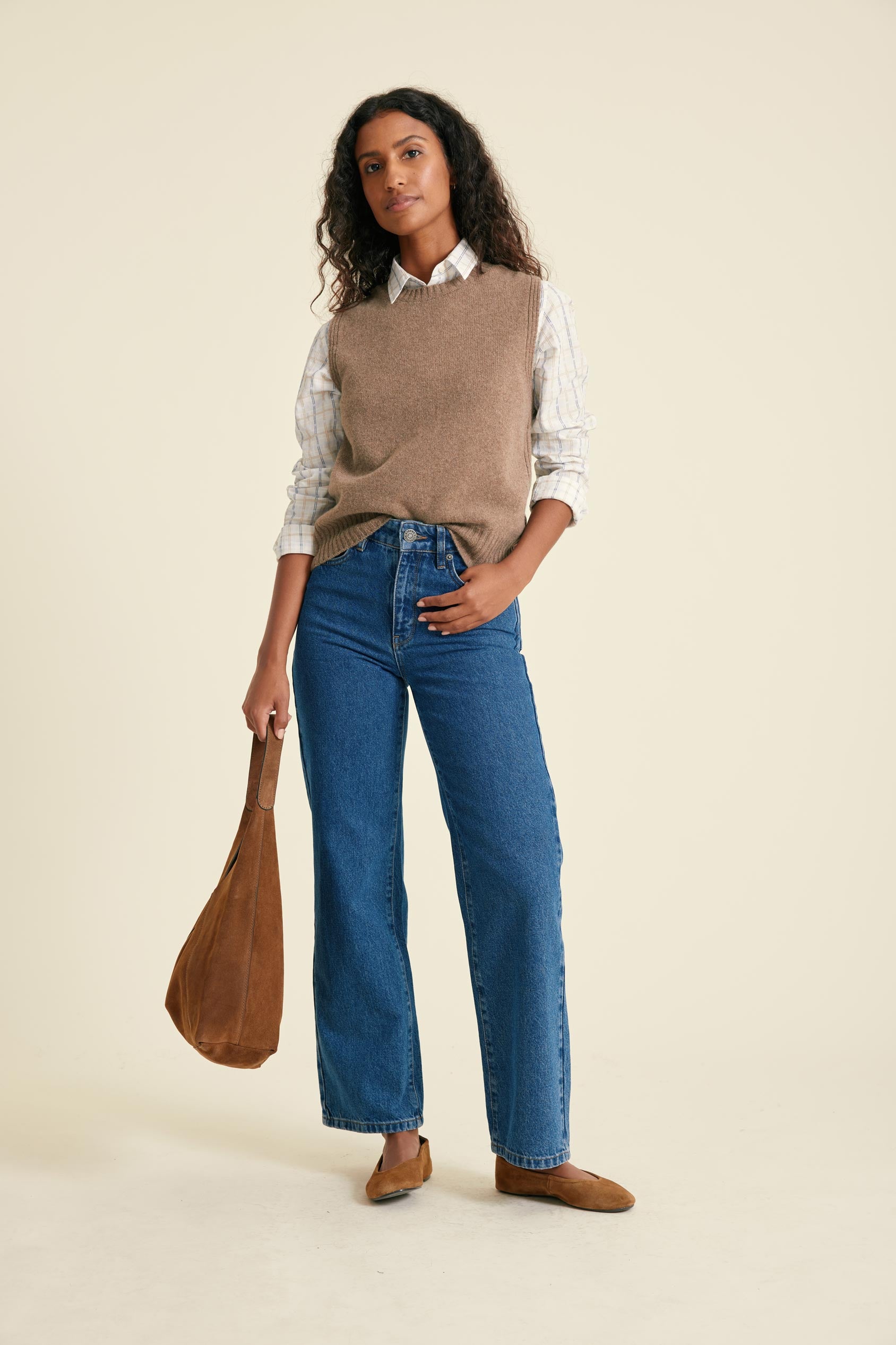 Woman wearing a beige sweater vest, white shirt, blue jeans, and brown shoes, holding a brown bag on a beige background.