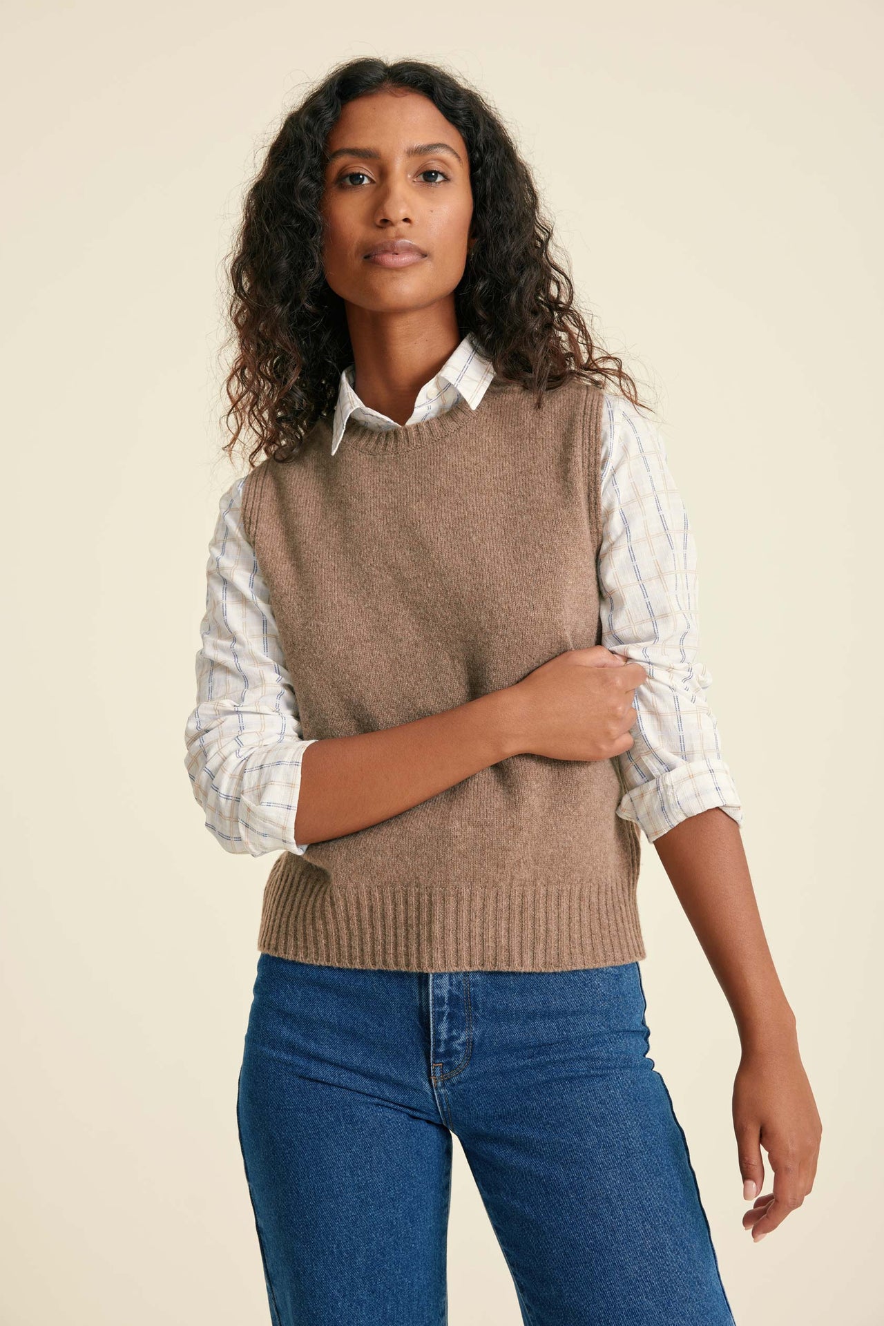 Woman wearing a brown sweater vest over a white shirt with rolled-up sleeves, paired with blue jeans on a beige background.