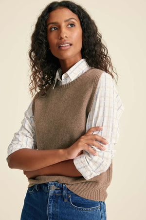 Woman wearing a brown sweater vest over a white shirt with rolled-up sleeves, standing against a plain background.