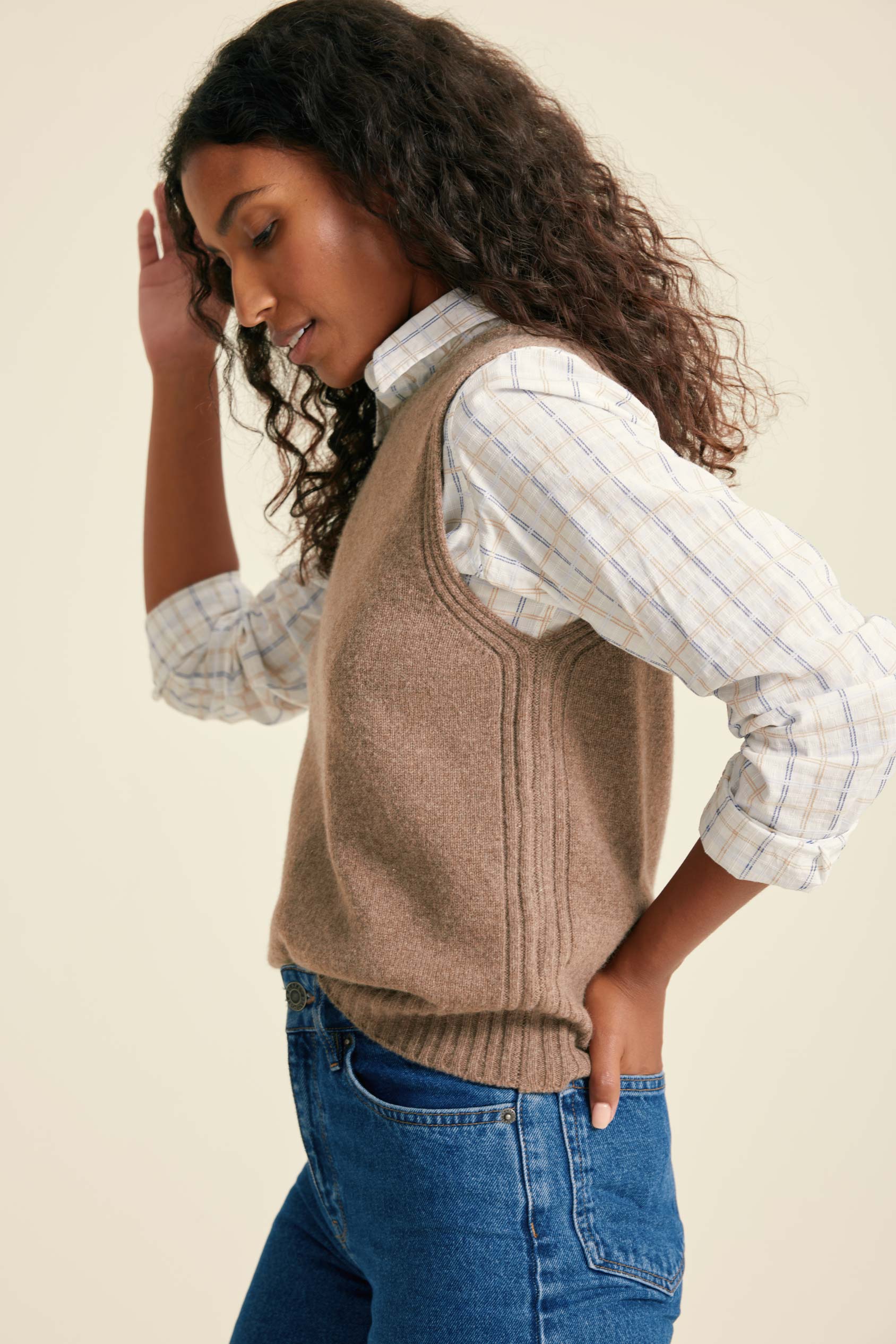 Woman wearing a beige sweater vest over a plaid shirt with rolled-up sleeves, standing against a plain background.