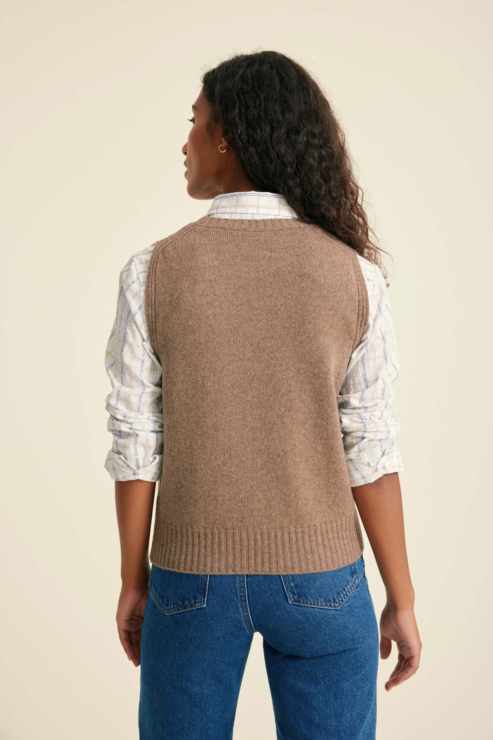 Person wearing a brown sweater vest over a patterned shirt with blue jeans on a beige background