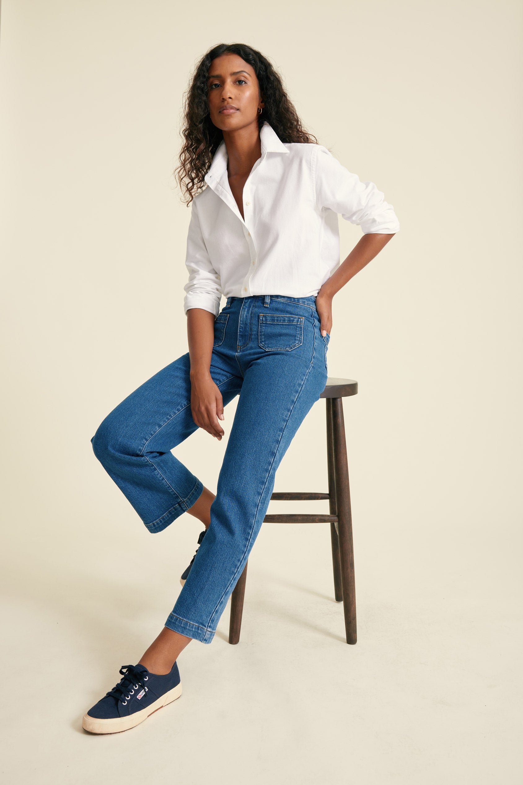 Woman wearing a white shirt and blue jeans sitting on a wooden stool against a beige background
