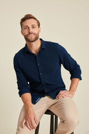 Man wearing a navy blue shirt and beige pants sitting on a stool against a plain background