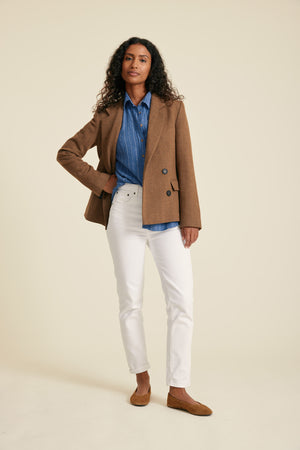Woman wearing a brown blazer, blue shirt, and white pants on a beige background