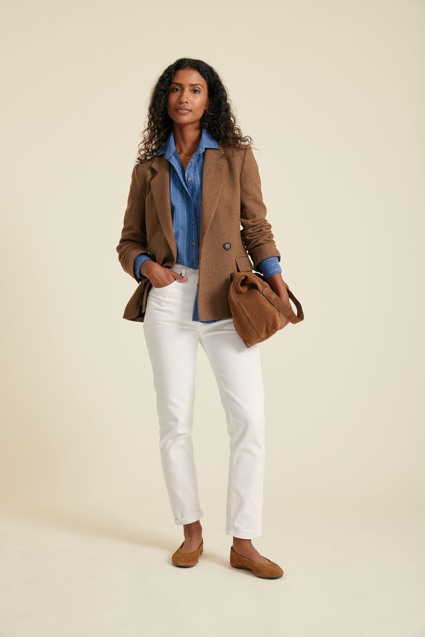 Woman wearing a brown coat, blue shirt, white pants, and brown shoes on a beige background