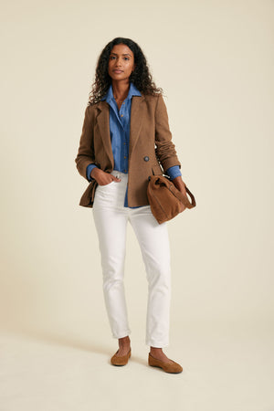 Woman wearing a brown coat, blue shirt, white pants, and brown shoes on a beige background