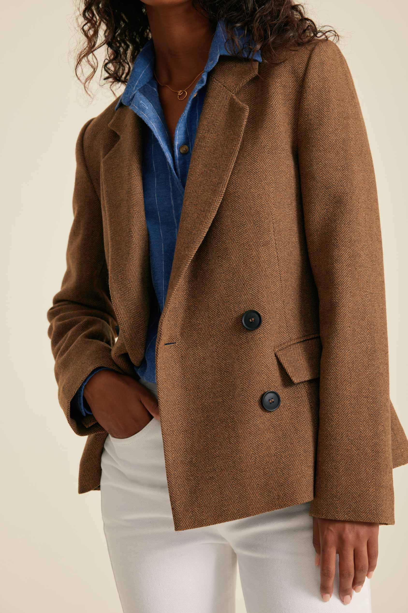 Person wearing a brown coat over a blue shirt and white pants on a beige background