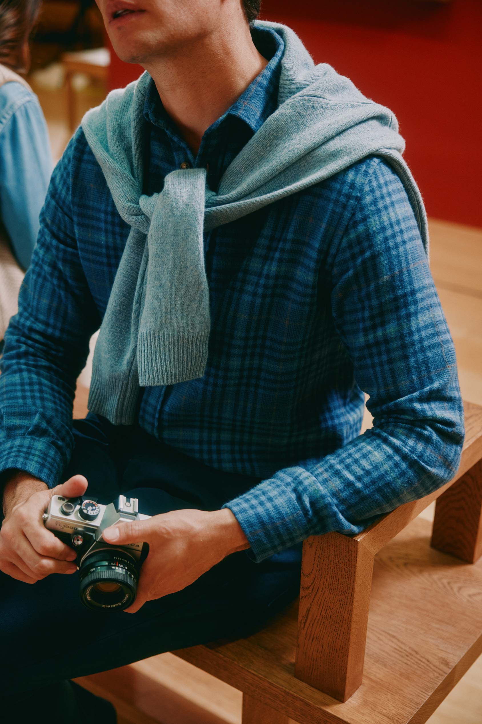 Person wearing a blue plaid shirt and light blue scarf, holding a camera indoors.