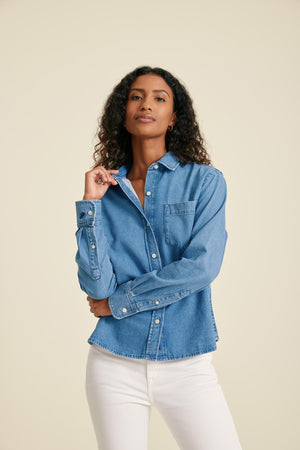 Woman wearing a blue denim shirt and white pants against a beige background