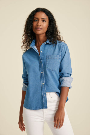 Woman wearing a blue denim shirt and white pants against a beige background