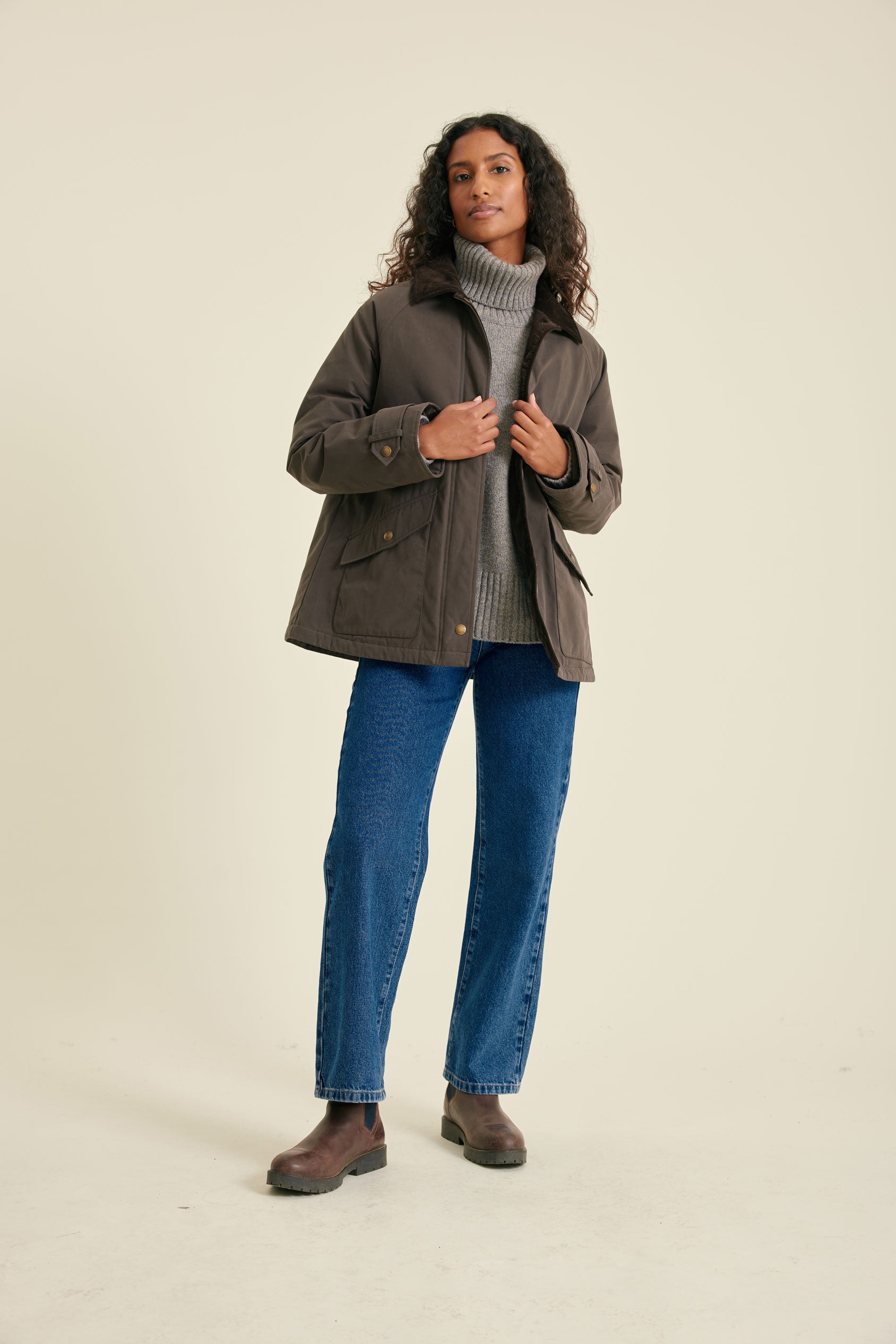 Woman wearing a brown jacket, gray sweater, blue jeans, and brown shoes on a beige background